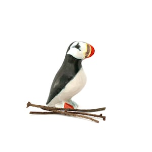 May include: A hand-painted wooden puffin figurine. The bird has a black back, white belly, and a distinctive orange and red beak. It is perched on a small, brown twig base against a white background. The puffin is a charming decorative item.
