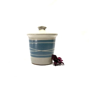 Peut inclure: Un pot en céramique blanche avec un motif rayé bleu et blanc et un couvercle. Le pot est posé sur une surface blanche.