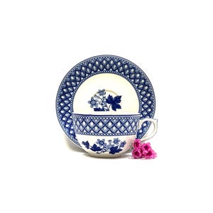 May include: A white teacup and saucer set with a blue floral and geometric pattern. The cup and saucer are placed on a white surface, with a small cluster of pink flowers to the side. The set is a classic design.