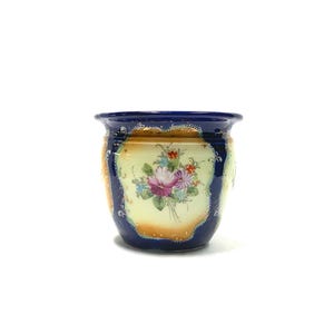 May include: A blue and gold ceramic planter with a floral design. The planter has a dark blue rim and accents, with a cream-coloured body adorned with a bouquet of pink, purple, and red flowers. The planter is a decorative item.