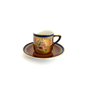 May include: A vintage teacup and saucer set. The cup and saucer are brown with a dark blue trim. The cup features a gold handle and a painted scene of people in a garden. The set is likely porcelain.