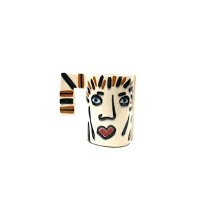 May include: White ceramic mug with a hand-painted face design. The face has black outlines, blue eyes, a red heart-shaped mouth, and brown and orange hair.