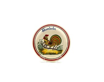 Puede incluir: Plato pequeño y redondo de cerámica con un diseño de gallo colorido. El gallo es amarillo, rojo y verde, de pie sobre hierba. El plato tiene un borde rojo y blanco, con la palabra "bhantecier" en azul.