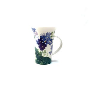 May include: A tall, white ceramic mug with a handle, adorned with a watercolor floral design. The design features blue and purple violets with green leaves. The mug is set against a white background.