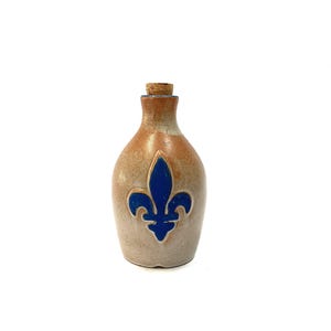 May include: A small, tan ceramic bottle with a cork stopper. The bottle has a blue fleur-de-lis design on the front. The bottle has a rounded shape and is suitable for storing liquids. The bottle is likely vintage or antique.