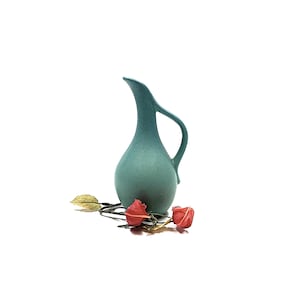 May include: A teal-colored ceramic pitcher with a curved handle and spout. Two dried red flowers and a gold-colored leaf are at the base of the pitcher. The background is white.