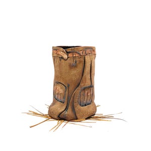 May include: A brown ceramic vase with a textured, organic design. The vase has carved details and a unique shape, sitting on dried grass. This handcrafted piece adds a rustic touch to home decor. A decorative item.