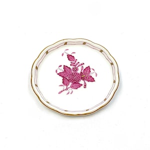May include: A small, round, white porcelain saucer featuring a scalloped edge with a thin gold rim. The center displays a detailed magenta floral motif with leaves and blossoms. A delicate magenta and gold striped pattern accents the scalloped rim.