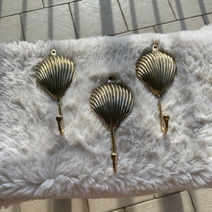 May include: Three gold-toned metal hooks shaped like seashells. The hooks are mounted on a white fluffy surface.