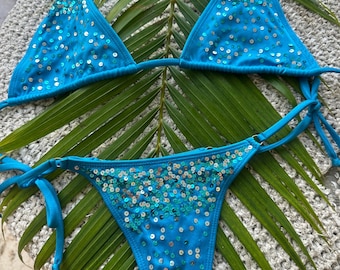 May include: A bright turquoise bikini set featuring a triangle top and tie-side bottoms. Both pieces are adorned with scattered, shimmering silver and teal sequins that create a sparkling effect across the fabric of the swimwear.