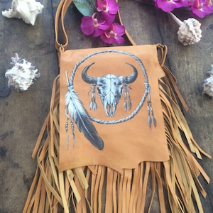 May include: A tan leather crossbody bag with a fringe trim. The bag features a painted design of a buffalo skull inside a circle with feathers and beads.