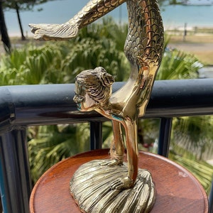 May include: A gold-toned mermaid figurine standing on a shell, with a tail and scales. The figurine is on a wooden pedestal.