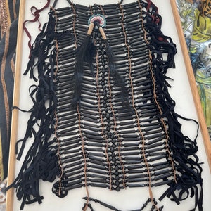 May include: A black beaded necklace with fringe and a small circular design in the center. The necklace is made of black beads and leather.