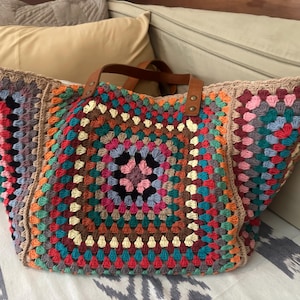 Puede incluir: Un bolso tote grande y colorido tejido a ganchillo con un diseño de cuadrados de la abuela. El bolso presenta una variedad de colores que incluyen rojo, azul, verde, naranja y marrón. Tiene asas de cuero marrón.