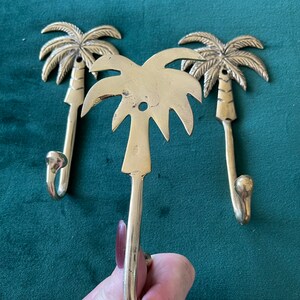 Puede incluir: Tres ganchos de oro en forma de palmera. Los ganchos est&aacute;n dise&ntilde;ados para parecerse a palmeras con un gancho curvo en la parte inferior. Los ganchos est&aacute;n sobre un fondo verde.