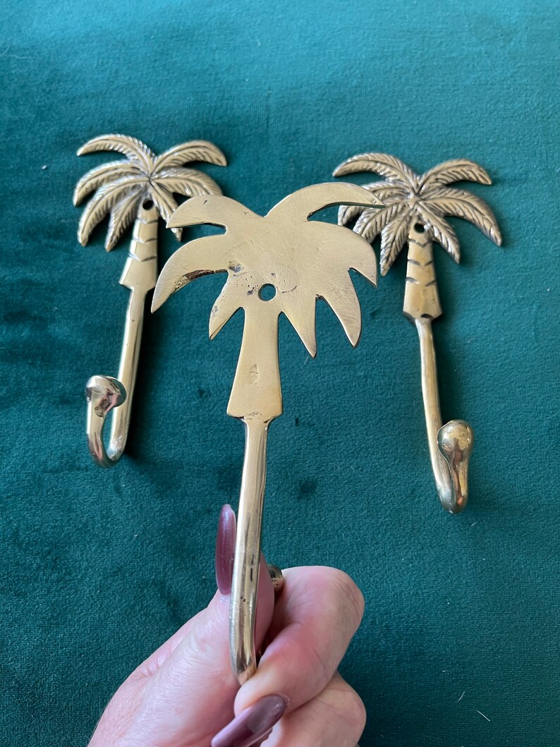 Puede incluir: Tres ganchos de oro en forma de palmera. Los ganchos est&aacute;n dise&ntilde;ados para parecerse a palmeras con un gancho curvo en la parte inferior. Los ganchos est&aacute;n sobre un fondo verde.