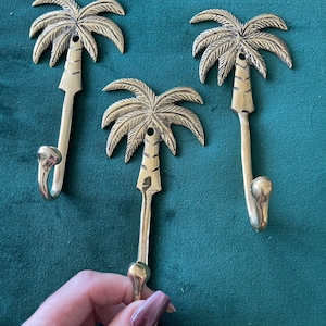May include: Three gold palm tree hooks. The hooks are shaped like palm trees with a curved hook at the bottom. The hooks are on a green background.