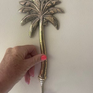 May include: A gold palm tree shaped hook for hanging items on a wall. The hook is made of metal and has a detailed design.