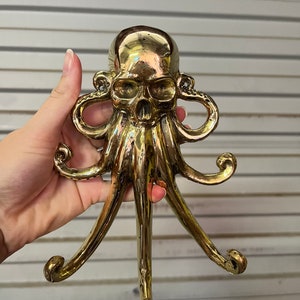 Puede incluir: Un perchero de color dorado en forma de pulpo con una cabeza de calavera. El pulpo tiene seis tentáculos que están curvados en forma de gancho.