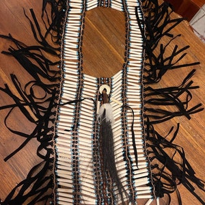 Puede incluir: Un collar de cuentas nativo americano con cuentas de hueso blanco, cuentas negras y detalles turquesa. El collar tiene una franja de cuero negro y una larga pluma negra y blanca que cuelga del centro.