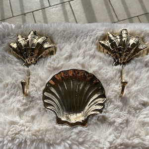 May include: Three gold-tone seashell hooks with a matching seashell dish. The hooks are designed to look like seashells and have a curved shape. The dish is a shallow, round dish with a scalloped edge.