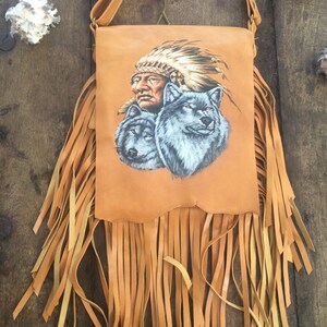 May include: A brown leather shoulder bag with fringe detailing. The bag features a painted design of a Native American man wearing a headdress and two wolves.