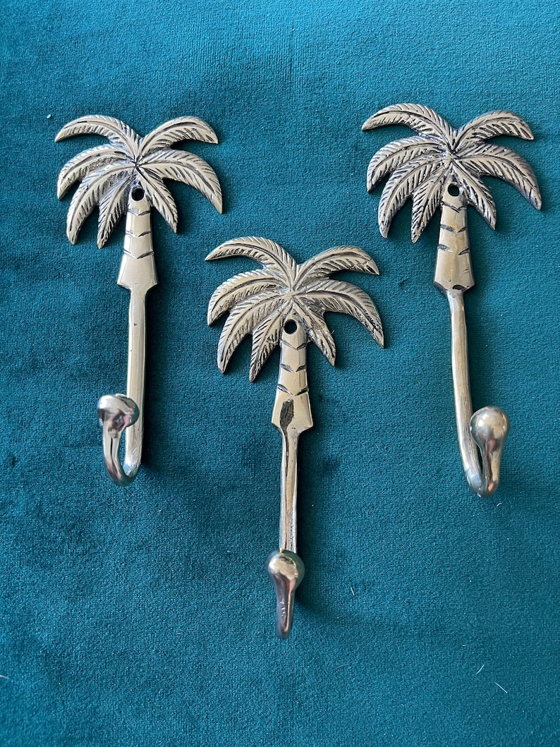 Puede incluir: Tres ganchos de palmera plateados. Los ganchos tienen forma de palmeras con un gancho curvo en la parte inferior. Los ganchos son una forma decorativa de colgar objetos en una pared.