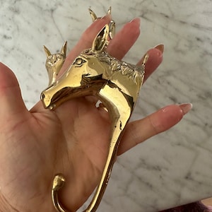 May include: A gold-toned metal horse head hook. The hook is shaped like a horse's head with a curved hook extending from the bottom.