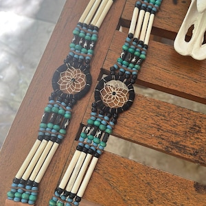 May include: Two handmade beaded bracelets with a dreamcatcher design. The bracelets feature cream-colored bone beads, turquoise, black, and blue beads, and a leather strap. The dreamcatcher is black with a tan web design.