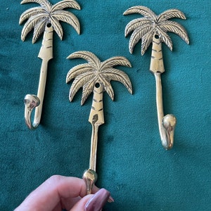 Puede incluir: Tres ganchos de palmera de oro. Los ganchos tienen forma de palmeras con un gancho curvo en la parte inferior. Los ganchos est&aacute;n sobre un fondo verde.