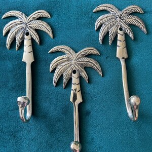 Puede incluir: Tres ganchos de palmera plateados. Los ganchos tienen forma de palmeras con un gancho curvo en la parte inferior. Los ganchos est&aacute;n sobre un fondo azul turquesa.