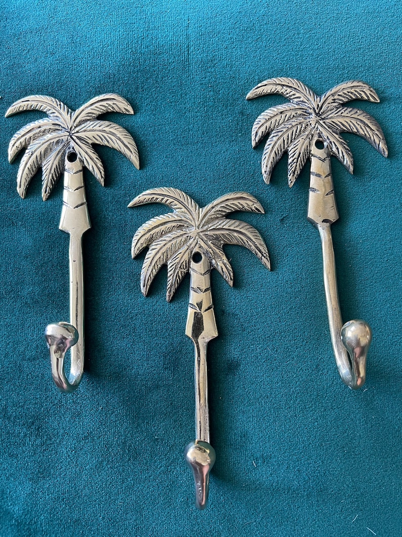 Puede incluir: Tres ganchos de palmera plateados. Los ganchos tienen forma de palmeras con un gancho curvo en la parte inferior. Los ganchos est&aacute;n sobre un fondo azul turquesa.