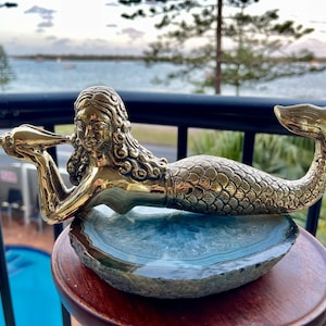 May include: A gold mermaid figurine holding a seashell, resting on a blue and grey agate slice. The figurine is detailed with scales and flowing hair.