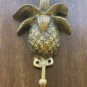 May include: A gold pineapple-shaped hook with a detailed design. The hook is mounted on a wooden surface.