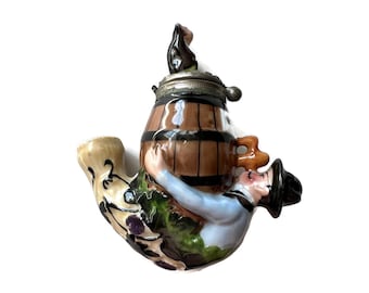 May include: A novelty ceramic pipe shaped like a barrel being embraced by a man in a blue shirt and black hat. The pipe has a silver lid and a curved stem with floral and grape designs.