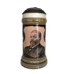 May include: A ceramic beer stein with a portrait of a man in a suit. The stein has a dark lid, a light-colored top, and a brown body with decorative bands. The base and rim have intricate designs.