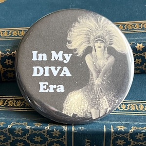May include: A black and white button with a vintage image of a woman wearing a feathered headdress and a sequined dress. The text "In My DIVA Era" is printed in white on the button.