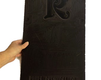 May include: A rectangular dark brown printing plate featuring a large stylized letter R at the top and the embossed text ROYCE CHEMICAL CO. EAST RUTHERFORD, NEW JERSEY at the bottom. The surface has a visible wood grain texture and small cross marks.