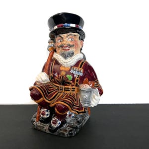 May include: A ceramic Toby jug figurine of a seated man in a burgundy and gold uniform, black top hat, and white collar. The jug has a handle and holds a small pewter mug. The figurine is on a black surface.