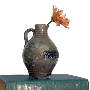 May include: A gray ceramic pitcher with a single handle and a narrow neck, featuring embossed floral patterns and a rectangular blue plaque with the text KOMM, TRINK ZUM WEIN. A single peach-colored flower is placed inside the pitcher opening.