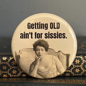 May include: A round button with a beige background and black text that reads "Getting OLD ain't for sissies."  A black and white photo of a woman sitting in a chair is in the background.