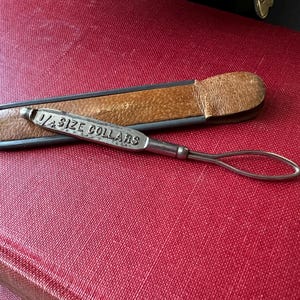 May include: A silver metal collar button tool with a brown leather case. The tool is engraved with the words "1/4 SIZE COLLARS".