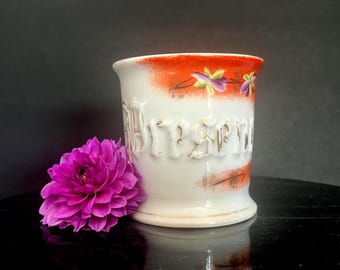 May include: A white ceramic mug featuring a raised, gold-lettered inscription that reads "Dresden." The upper rim is decorated with a vibrant orange band and hand-painted purple and yellow floral accents. A bright magenta dahlia flower sits beside it.