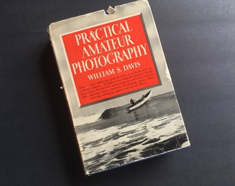 May include: A vintage book titled "Practical Amateur Photography" by William S. Davis. The cover features a red rectangle with white text, and a black and white illustration of a boat on the water. The book is worn, with visible signs of age.