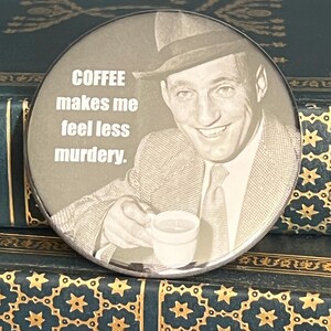 May include: A round, sepia-toned button featuring a smiling man in a fedora holding a coffee cup. The text "COFFEE makes me feel less murdery" is printed in white. The button is set against a background of antique books.