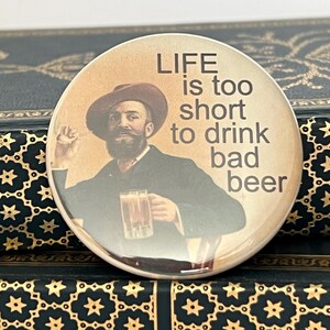 May include: A round button with a vintage illustration of a man in a hat holding a beer mug. The button has the text "LIFE is too short to drink bad beer" in a serif font. The background is a sepia tone.