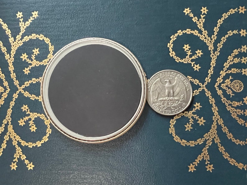 May include: A round, silver-rimmed magnet with a black center, next to a U.S. quarter coin. The magnet is on a dark teal surface with gold floral designs. The quarter shows an eagle and the words "Quarter Dollar."