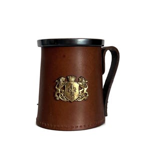 May include: A brown leather mug with a black rim and handle. A gold-colored emblem with a crown, fleur-de-lis, and lions is affixed to the front. The mug has a classic, rustic appearance.