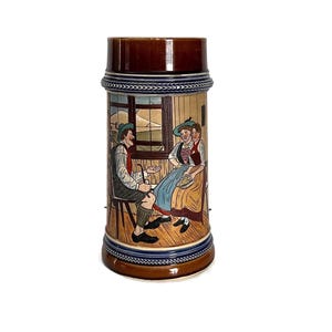 May include: A brown ceramic beer stein with a detailed painted scene of two people in traditional clothing. The stein has a dark brown lid and base, with blue and white decorative bands. The scene depicts a man and woman in a room.