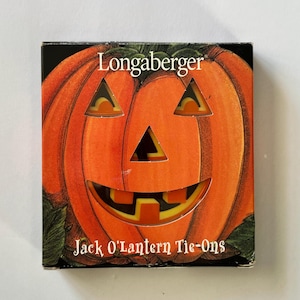 May include: A square product box featuring a large orange pumpkin illustration with a carved jack-o'-lantern face. The text Longaberger is printed at the top, and Jack O'Lantern Tie-Ons is printed at the bottom in white serif lettering.